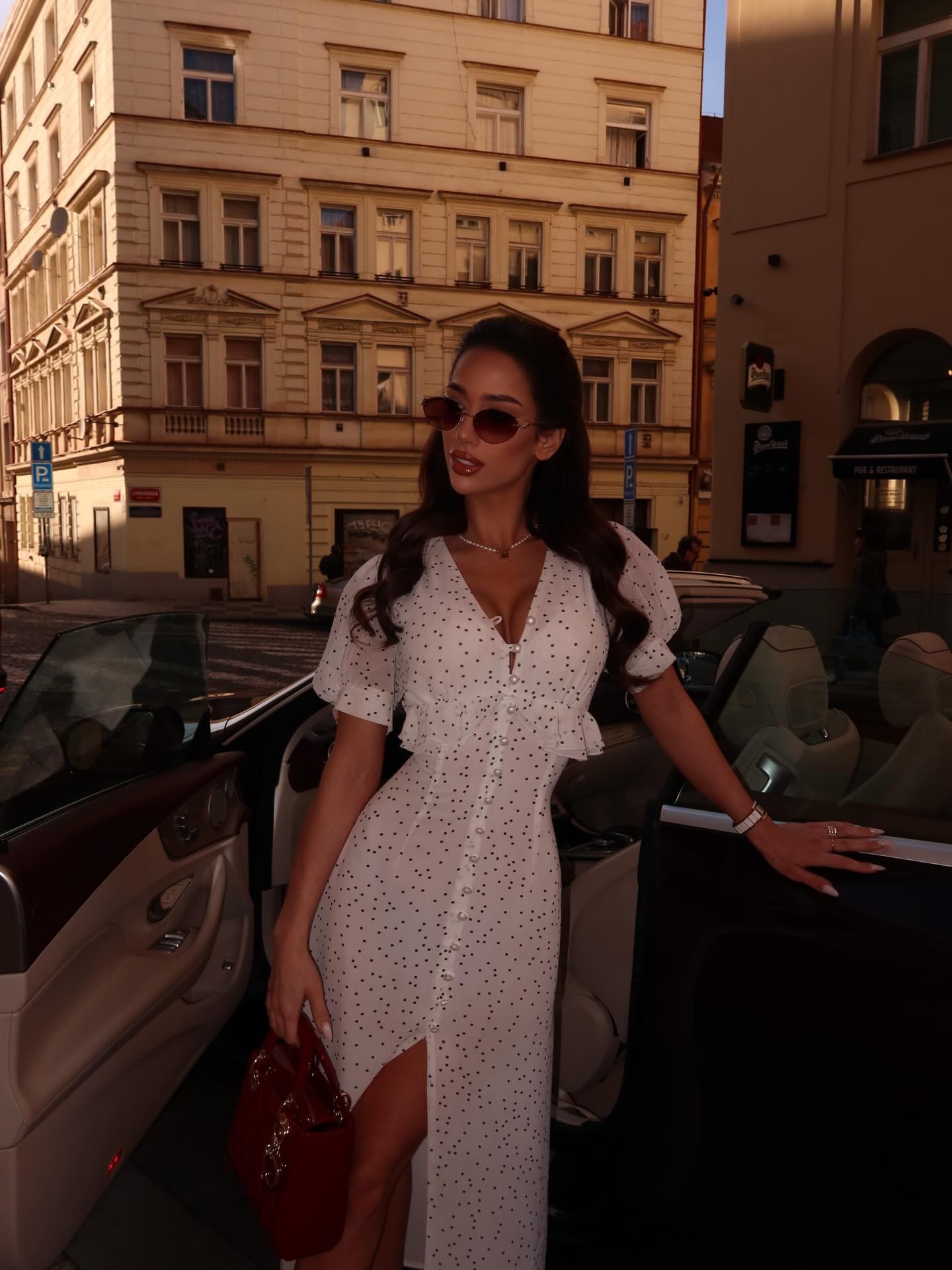 A woman stands beside an open car door in a white polka-dot dress with a high slit, holding a red bag.